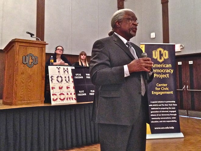 Frank Thompson in a suit holds a microphone in front of an auditorium