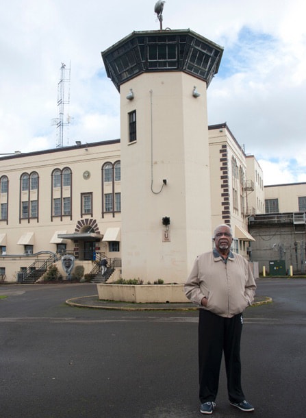 An older Frank Thompson wearing a track jacket stands outside near a penitentiary watch tower.