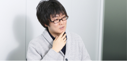カーディガンを着た眼鏡の男性が椅子に座り、あごに手をあて考えながら話している。