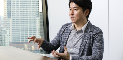 高層ビルを見通す窓のそばに座る男性が、手振りを交えて話している。
