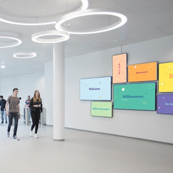 Employés Trivago qui marchent devant un mur d’écrans de télévision où apparaît le mot « Bienvenue » en plusieurs langues