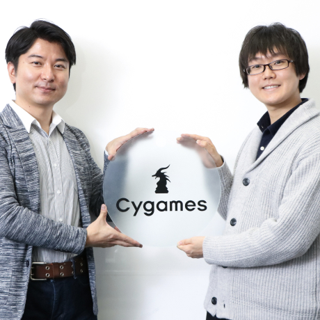 Cygames のロゴが刻まれた丸いガラスのサインプレートを挟んで微笑む二人の男性