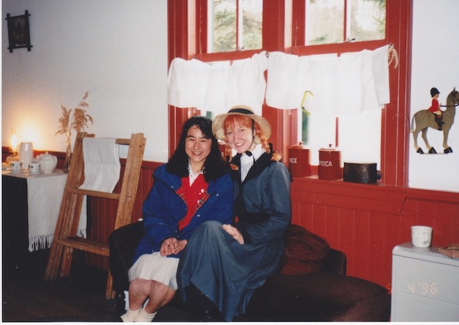 Erin wears a orange yarn wig and hat and poses with a coworker in a room with red trim and a Canadian Mountie decoration.