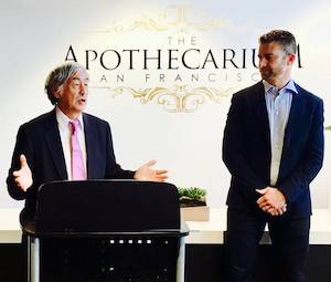 Two men in suits stand in front of an ornate logo for The Apothecarium.