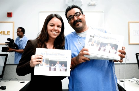 Kathryn Hymes, bénévole Slack, et une personne incarcérée sourient côte à côte, chacun tenant dans les mains un exemplaire du San Quentin News.